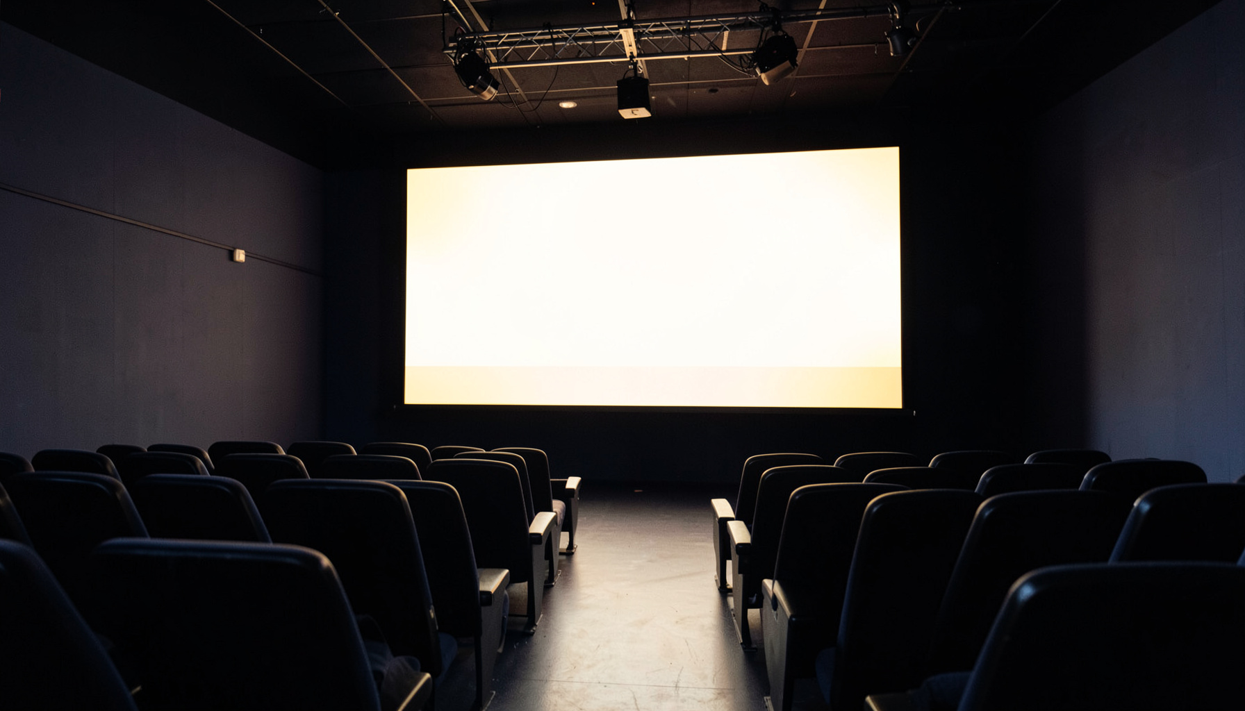 A darkened screening room with rows of empty seats facing a large glowing screen, warm amber and deep navy tones