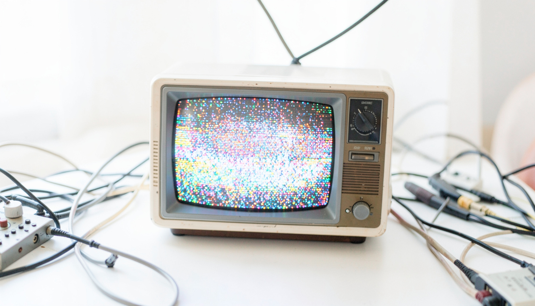 A bright, airy photograph of a vintage television set on a plain surface, its screen glowing with abstract colorful static, surrounded by scattered broadcast equipment cables